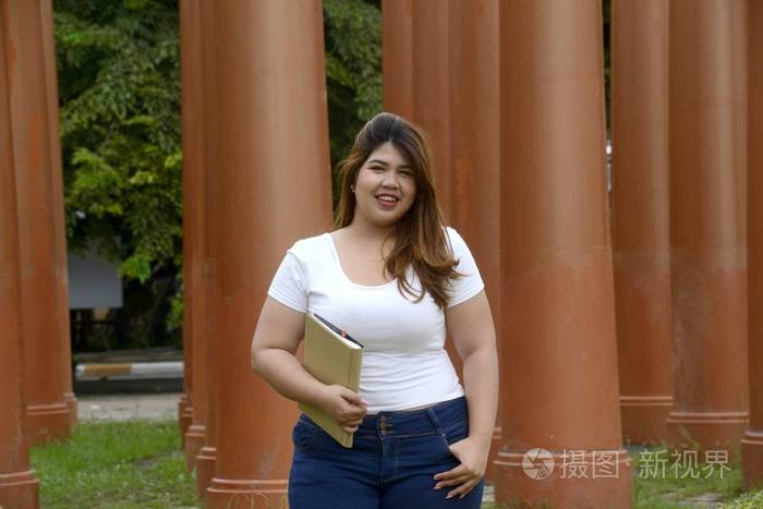 亚洲漂亮笑脸脸胖女人的肖像站在公园里的柱子上拿着一本小册子照片