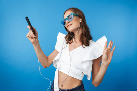 一个年轻女孩的肖像在夏天的衣服戴墨镜听音乐与耳机和手机在蓝色背景下隔离