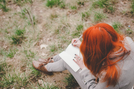 红头发的女孩穿着灰色的大衣坐在绿色的春天的草地上