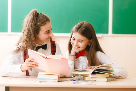 2女生在学校制服坐在一个绿色板的背景在书桌上的书. 姐姐教年轻读