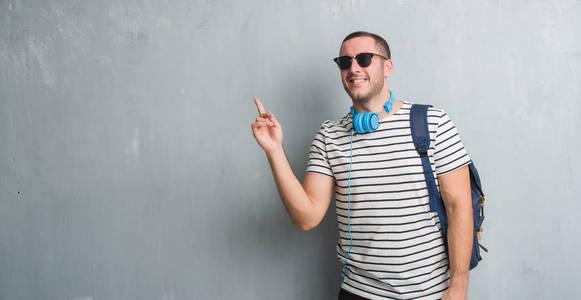 年轻的高加索学生男子在灰色的垃圾墙上戴着耳机和背包非常高兴地指着手和手指的侧面