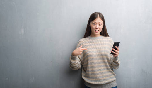 灰色墙壁的年轻中国妇女看在智能手机与惊奇面孔指向自己手指