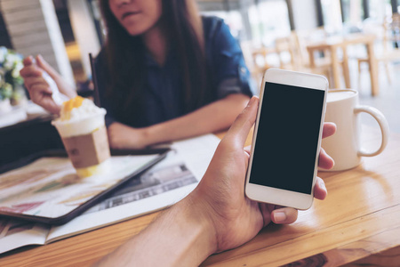 在现代咖啡馆和背景模糊的女人手中手持白色手机与空白黑屏的人的样机图像