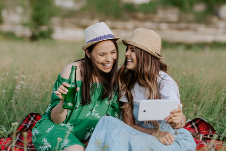 两个漂亮的年轻女人坐在野餐毯上, 玩得开心。啤酒和三明治