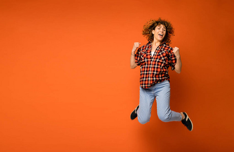 兴奋卷曲的女人在格子衬衫和牛仔裤站在橙色背景与空白的文字旁边她跳的幸福