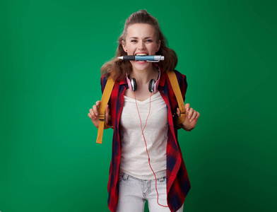 开朗的年轻学生妇女在红色衬衫与背包和耳机持有大笔在牙齿上的绿色背景