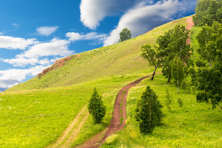 在阳光明媚的日子里, 在明亮的蓝天和云雾的背景下, 在森林边的山坡上, 用空旷的尘土飞扬的土路与夏日的风景