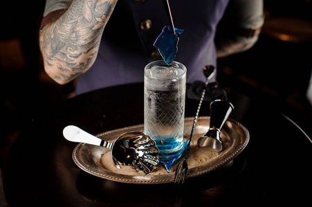 酒保与纹身制作新鲜和甜美的夏季鸡尾酒与蓝色的焦糖在托盘与沙子。海运风格