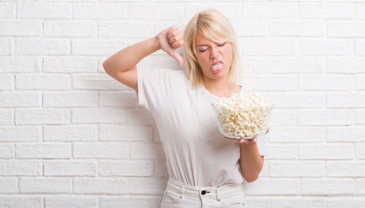 成年白人妇女在白色砖墙吃流行玉米与愤怒的面孔, 消极标志显示不喜欢以拇指下来, 拒绝概念