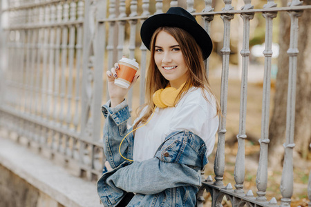 照片中, 一个美丽的微笑的嬉皮士女孩棕色头发戴着帽子, 拿着一杯茶或咖啡在公园里