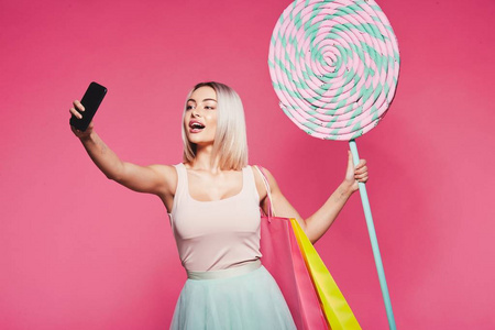 美丽的年轻女孩拿着五颜六色的购物袋和大棒糖采取自拍智能手机站在粉红色背景, 复制空间
