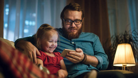 晚上爸爸和小女儿坐在客厅里看沙发上对智能手机的搞笑视频