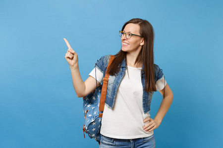 年轻漂亮女学生的肖像在牛仔服装眼镜与背包看一边指着在蓝色背景上孤立的复制空间。中学大学的教育理念