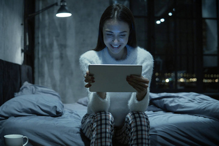 年轻的微笑的妇女坐在她的床上与数字平板电脑和看电影或网上购物深夜