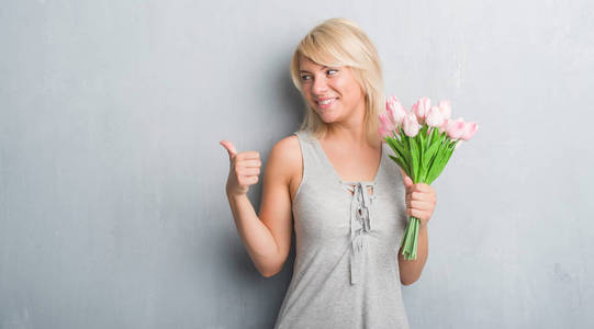 白人成年妇女在灰色的垃圾墙上举行粉红色的花朵指向和显示与拇指的一面微笑着快乐的脸