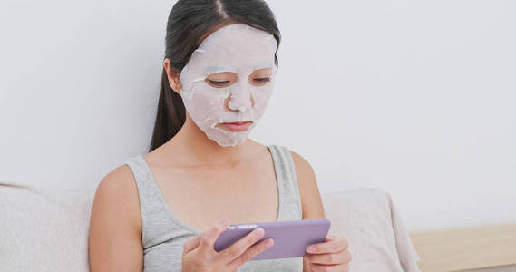 妇女在床上应用纸口罩和使用手机