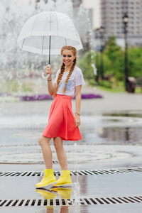 年轻漂亮的女孩与两个辫子在黄色靴子和透明的雨伞站在喷泉附近。城市的雨天