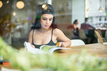 一个年轻美丽的女孩独自坐在咖啡厅和阅读时尚杂志的肖像