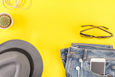 平躺在蓝色牛仔裤, 灰色帽子, 仙人掌和智能手机耳机在大胆的黄色纸背景与复制空间。女性休闲服装的头顶景色。时髦的时髦的看顶部视图