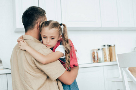父亲和伤心的小女儿背着背包拥抱在家里的厨房, 回到学校概念