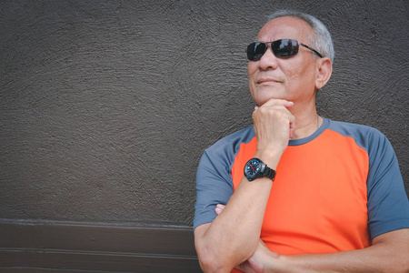 一位体贴的老人戴着太阳镜站在棕色墙边。亚洲老年男性的思想。高级休闲生活方式