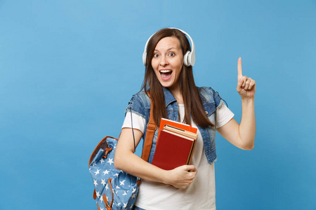 年轻惊奇女学生与背包耳机听音乐举行学校书指向索引手指在拷贝空间被隔绝在蓝色背景。高中院校教育