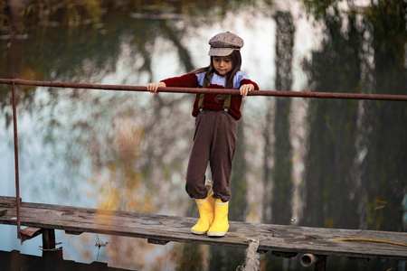 戴帽子和黄色橡胶靴的女孩站在河的背景木桥上