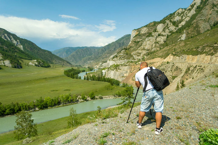 年轻男子摄影师通过阿尔泰与相机在三脚架上拍摄的积雪覆盖的山脉和岩石与绿松石缠绕河 Katun。照片浏览