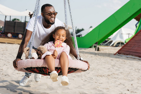 非洲裔美国人父亲推女儿蜘蛛网巢摇摆与棒糖在游乐园