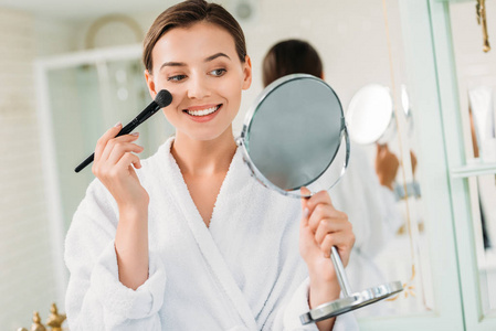 快乐的年轻女子浴袍手持镜和化妆在浴室