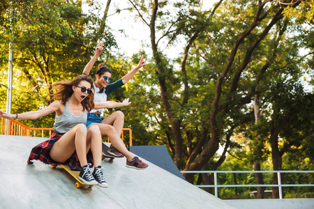 两个兴奋的年轻女孩有乐趣, 而骑滑板在公园的坡道