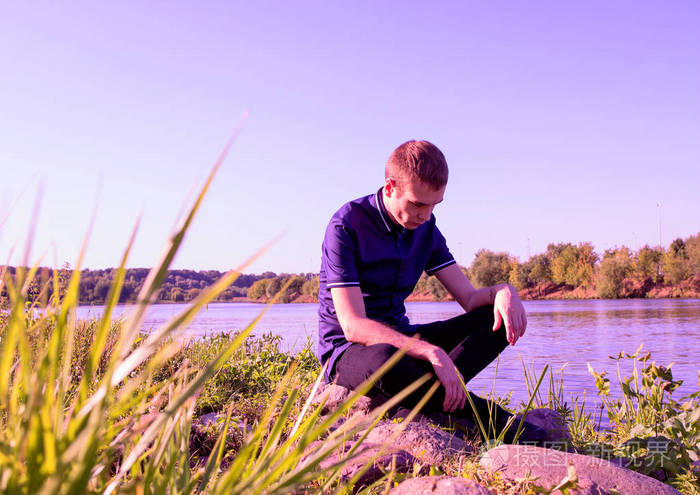 一个悲伤的年轻瘦家伙穿着蓝色衬衫 坐在河边的石头上 向下看照片