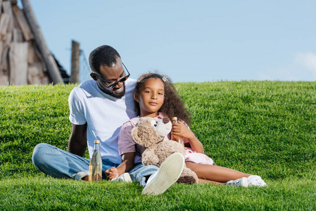 非洲裔美国人的父亲和女儿坐在山上与苏打和泰迪熊在游乐园里