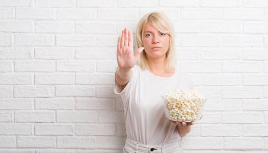 成年白人妇女在白色砖墙吃流行玉米用张开手做停止标志以严肃和自信的表达, 防御手势