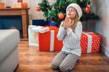 小女孩坐在她的膝盖上祈祷。她闭上眼睛, 手牵着手。希望和说谢谢的孩子。她身后有礼物的盒子和圣诞树