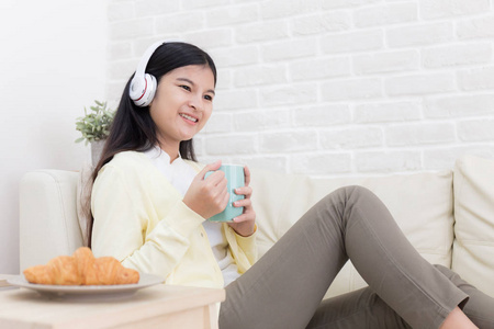 亚洲妇女正在听音乐从耳机和拿着一杯咖啡在家里的沙发上