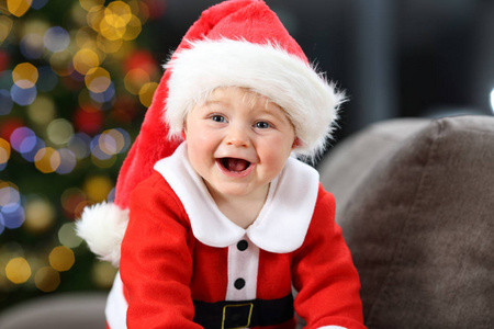 快乐的婴孩穿着红色的圣诞老人粘土伪装在沙发上在家里在圣诞节与树在背景