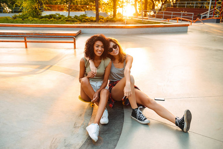 两个微笑的年轻女孩坐在公园里玩滑板的乐趣