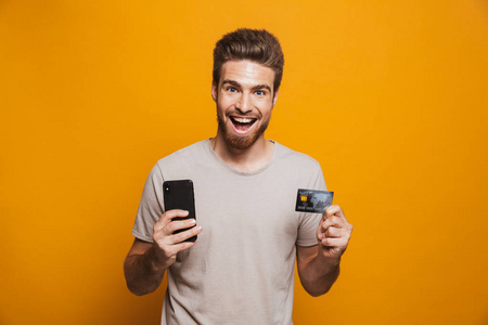 一个快乐的年轻人手持手机和显示在黄色背景下隔离信用卡的肖像
