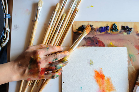 女性艺术家的手在凌乱肮脏的调色板与不同的油漆和画笔在艺术工作室拍摄从顶端点。白色背景