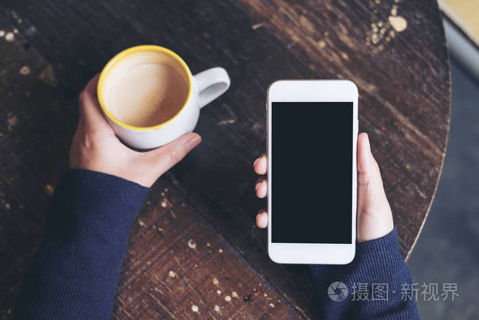 样机顶部视图女性手拿着白色的手机与空白的黑屏和咖啡杯在老式咖啡馆
