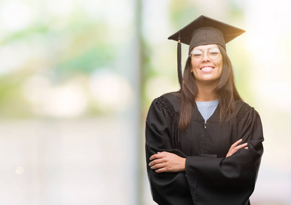 年轻的西班牙裔妇女穿着毕业帽和均匀快乐的脸微笑着交叉双臂看着相机。积极的人