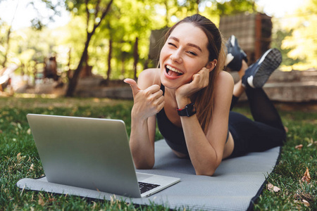 一个微笑的年轻健身女孩的肖像使用笔记本电脑, 而铺设在户外垫子上, 并显示竖起大拇指