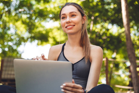 一个微笑的年轻健身女孩的肖像使用笔记本电脑, 而坐在户外