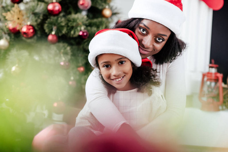 非洲裔美国人母亲在圣诞老人帽子拥抱小女儿在装饰房间在家里