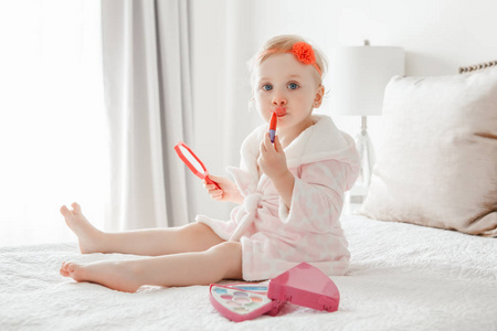 可爱的 asorable 滑稽的白种金发女孩2岁, 穿着浴衣做化妆, 看着镜子, 坐在她的床上在家里的卧室。快乐童年概念