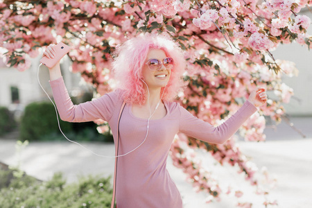 迷人的年轻女子与卷曲的粉红色的头发和太阳镜附近盛开的春天树听音乐智能手机