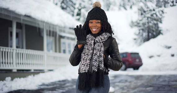 可爱的黑人妇女在舒适的帽子和围巾站在外面的房子在雪的挥舞