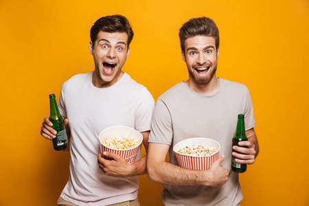 两个兴奋的情绪英俊的男人朋友站在黄色的墙上背景喝啤酒吃爆米花看相机