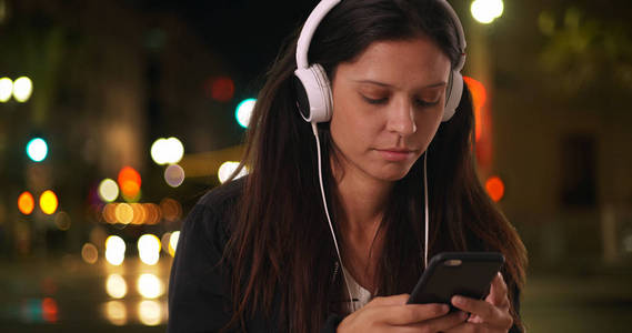 千禧年的女人在晚上在城市街道上听音乐时通过电话发短信